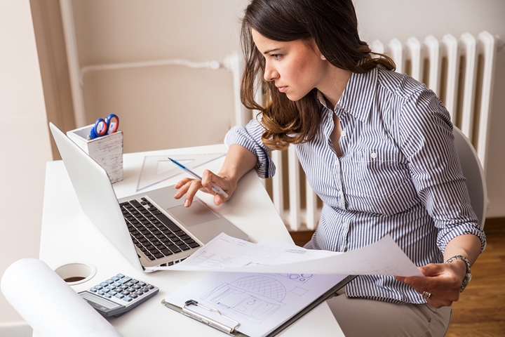 Eine Frau sitzt beschäftigt am Schreibtisch, wo sie Mitschriften mit einem Arbeitsdokument am Laptop abgleicht. 