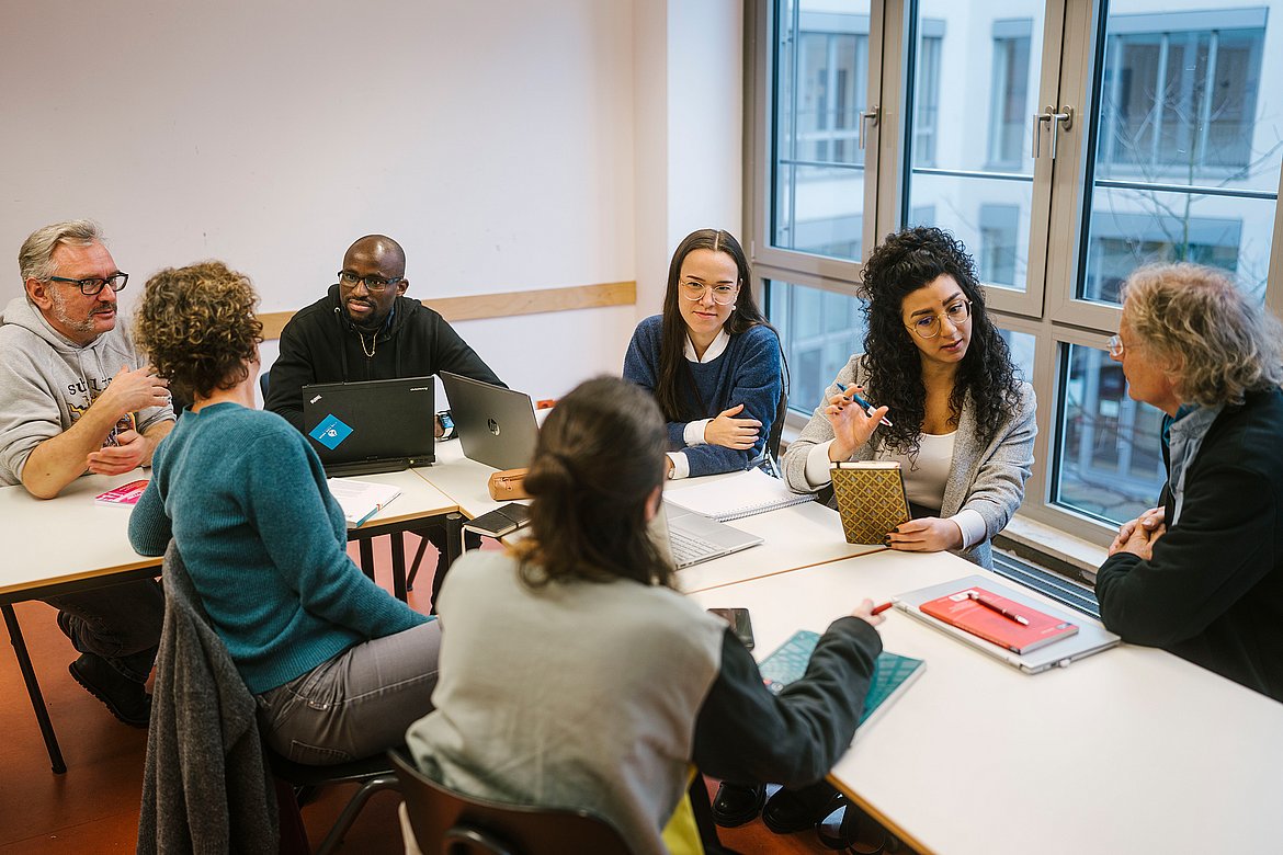 Studierende der ASH Berlin sitzen in einem Seminarraum nebeneinander sn einem Tisch und unterhalten sich. 
