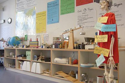 Blick in die Lernwerkstatt, Regale mit Experimentiergeräten, auf der rechten Seite ein Skelett mit Mantel und Schirmmütze.