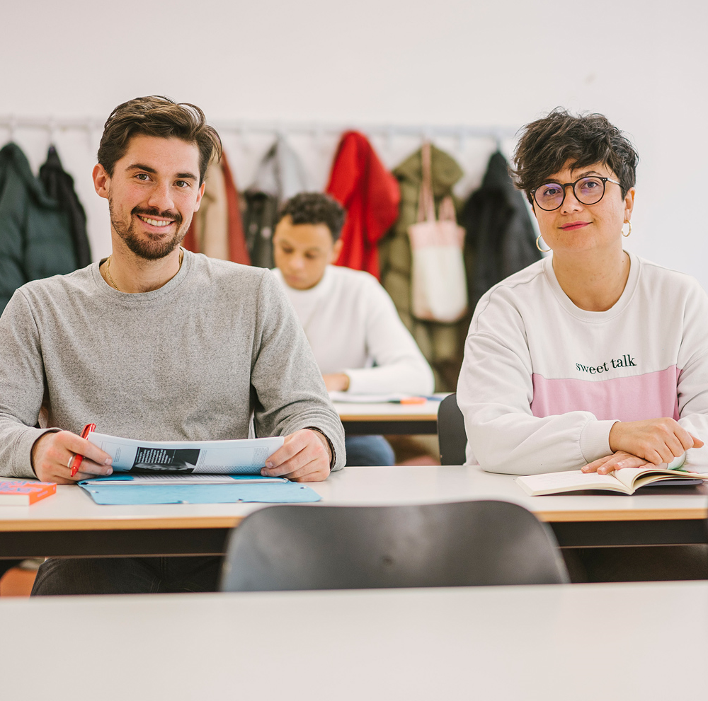 Auf dem Foto sind zwei Studierende der ASH Berlin zu sehen. Sie sitzen an einem Tisch in einem Seminarraum und schauen den Betrachter_innen in die Augen.