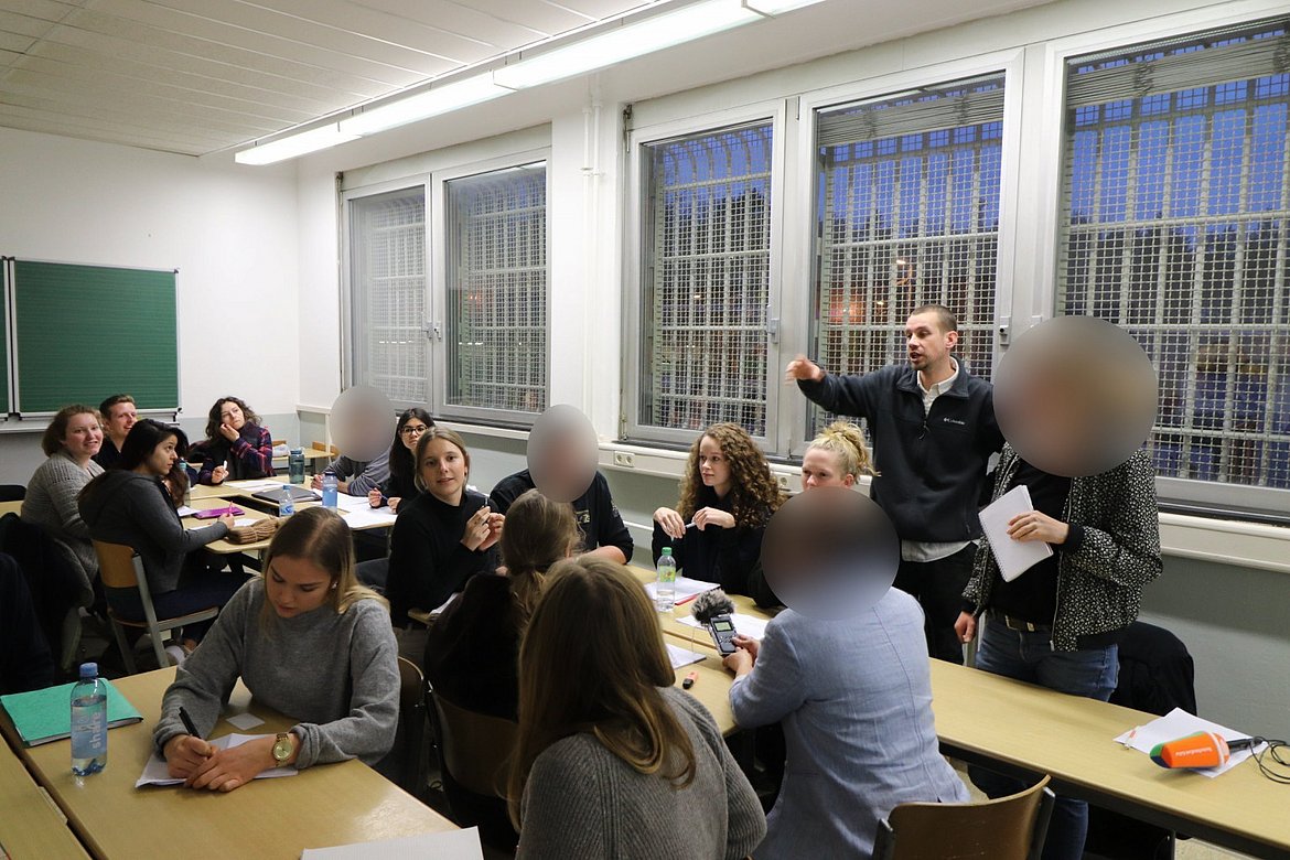 Abgebildet wird ein Seminarraum in einem Gefängnis, der Dozent Julian Knop steht und spricht zu den im Raum sitzenden Studierenden. 