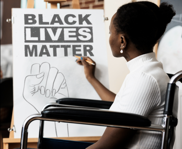 Eine schwarze Junge Frau sitzt im Rollstuhl und malt Black Lives Matter