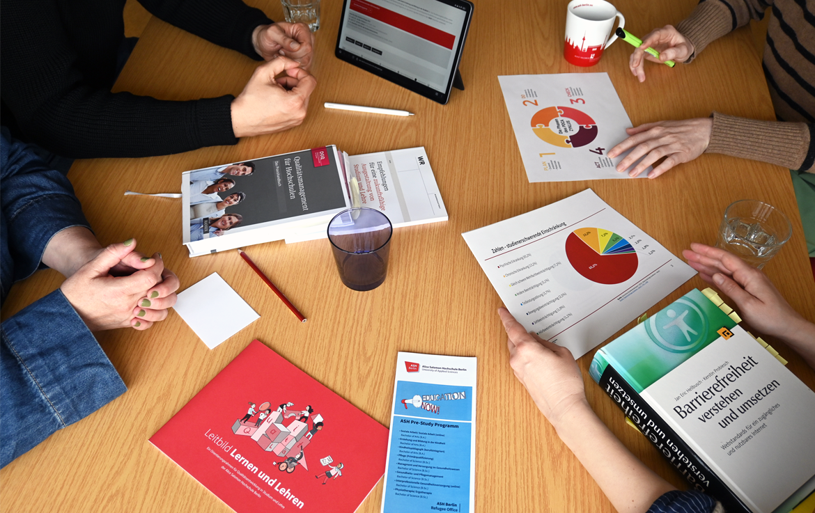 Tisch mit 4 Personen, von denen nur die Hände sichtbar sind, mit verschiedenen Ausdrucken, Broschüren, Faltblättern, Büchern zum Themenfeld Qualitätsentwicklung