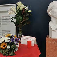The photo shows a table on which there are flowers, a candle and a card with text. The text reads, "In memory of all people affected by terror, war and oppression." Next to the table is a bust of Alice Salomon. 