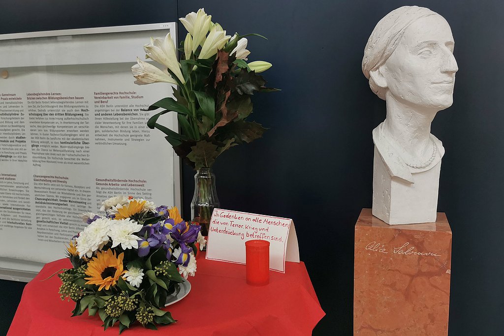 The photo shows a table on which there are flowers, a candle and a card with text. The text reads, "In memory of all people affected by terror, war and oppression." Next to the table is a bust of Alice Salomon. 