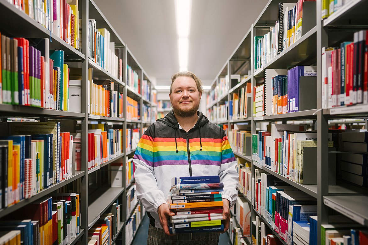 Eine Person mit Regenbogenjacke steht zwischen den Bücherregalen der Bibliothek und trägt einen Stapel Bücher in der Hand