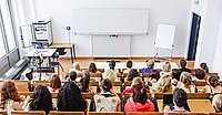 Blick von hinten über einen Hörsaal, Studierende sitzen in den Rängen, vorne eine leere Tafel