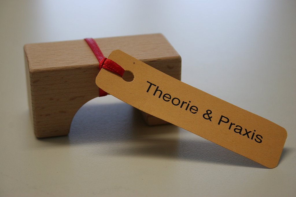 Ein kleiner Holzklotz in der Form ähnlich wie eine Bogenbrücke mit einem Zettel dran gebunden auf dem Theorie und Praxis steht