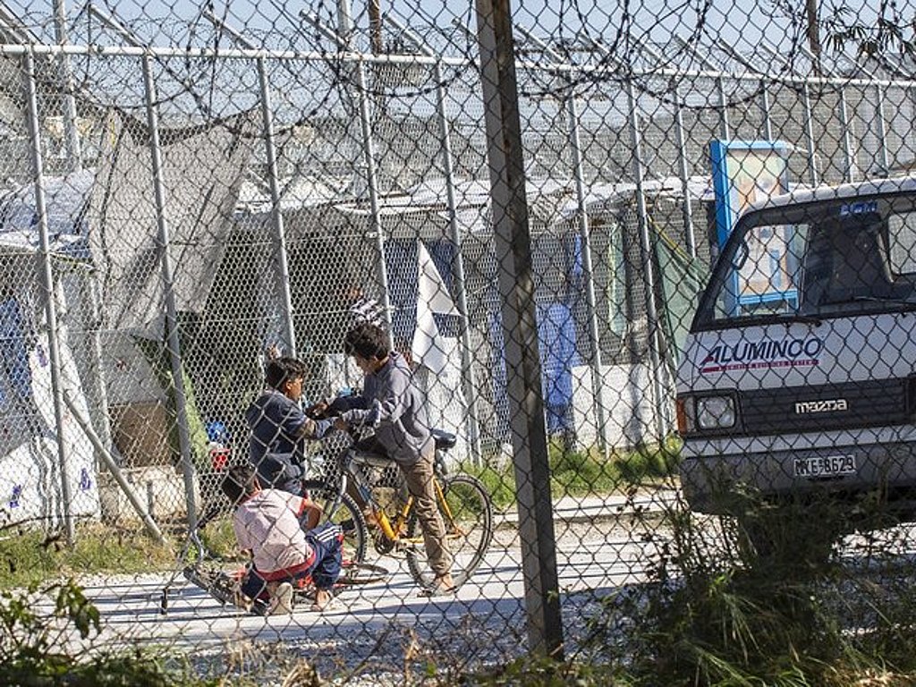 Blick durch ein eingezäuntes Camp in Griechenland - im Zentrum sieht man drei Kinder um ein Fahrrad, dahinter erkennt man noch mehr Zaun und Strukturen von behelfsmäßiger Unterbringung