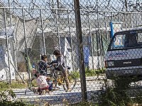 Blick durch ein eingezäuntes Camp in Griechenland - im Zentrum sieht man drei Kinder um ein Fahrrad, dahinter erkennt man noch mehr Zaun und Strukturen von behelfsmäßiger Unterbringung
