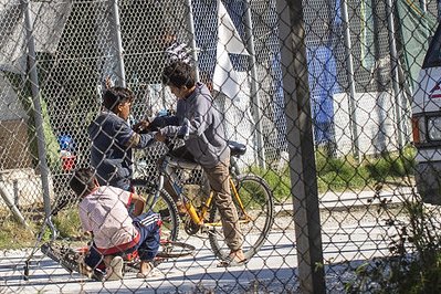 Blick durch ein eingezäuntes Camp in Griechenland - im Zentrum sieht man drei Kinder um ein Fahrrad, dahinter erkennt man noch mehr Zaun und Strukturen von behelfsmäßiger Unterbringung