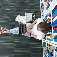 Person sitzt in einer Bibliothek auf dem Fußboden mit einem Laptop auf dem Schoß und arbeitet