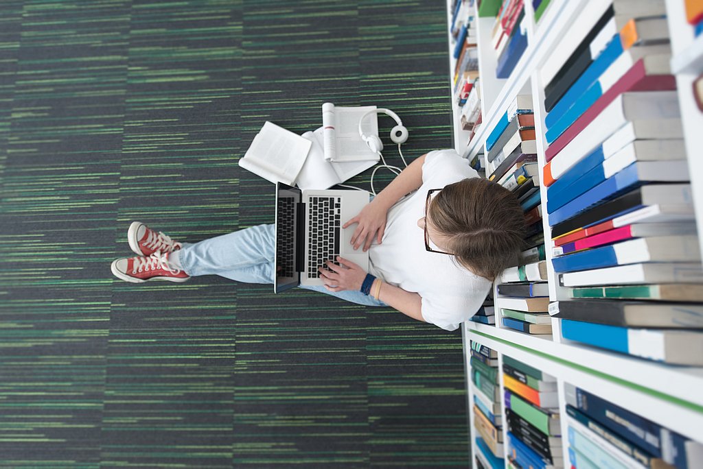 Person sitzt in einer Bibliothek auf dem Fußboden mit einem Laptop auf dem Schoß und arbeitet