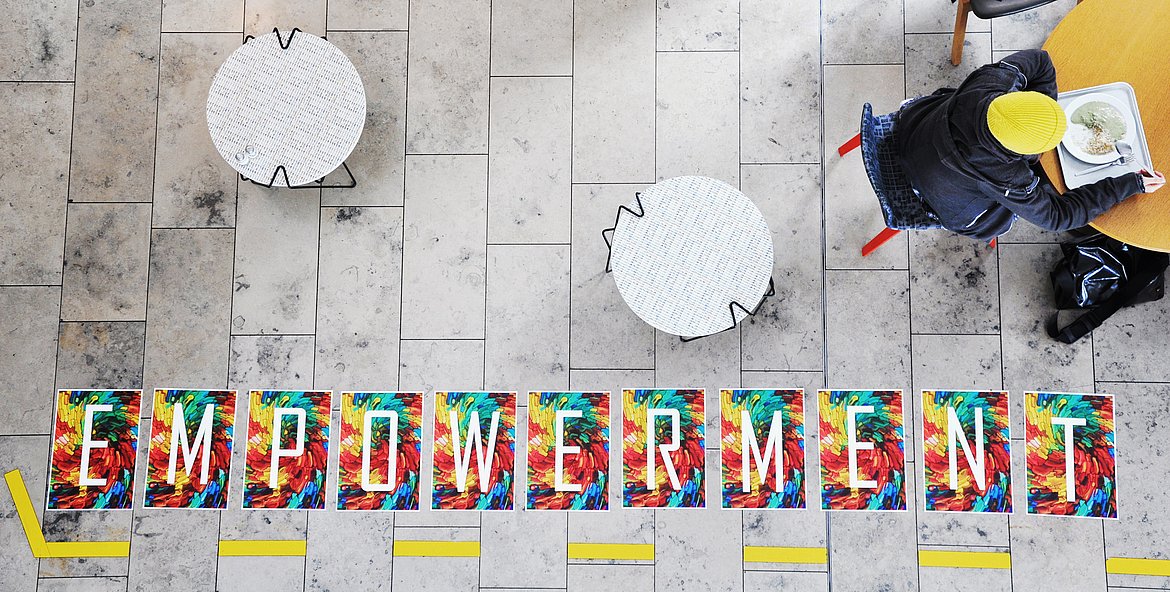 [Translate to Englisch:] Wort "Empowerment" auf dem Boden in der Mensa. Jemand sitzt und isst