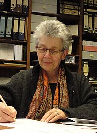The photo shows Adriane Feustel sitting at a desk and working. In the background you can see bookshelves. 