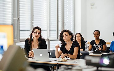 Mit Laptops ausgestattete Studierende des Online Bachelor Studiengangs Soziale Arbeit blicken gespannt nach vorne, wo ein Beamer eine Präsentation auf die Wand wirft