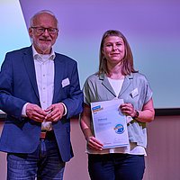 Auf dem Foto sind 2 Personen zu sehen die nebeneinander stehen und in die Kamera lächeln. Sie halten ein Geschenk bzw. eine Urkunde in der Hand.