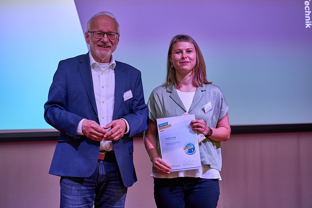 Auf dem Foto sind 2 Personen zu sehen die nebeneinander stehen und in die Kamera lächeln. Sie halten ein Geschenk bzw. eine Urkunde in der Hand.