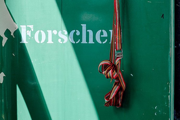 Detailaufnahme: an einer grünen Wand mit abblätternder Farbe baumelt von der leeren, orangenen Halterung eines Verbandkastens ein Spanngurt. Auf der Wand steht: Forschen