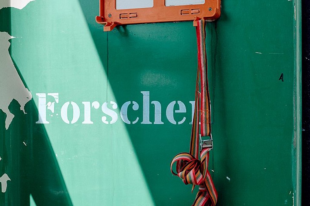 Detailaufnahme: an einer grünen Wand mit abblätternder Farbe baumelt von der leeren, orangenen Halterung eines Verbandkastens ein Spanngurt. Auf der Wand steht: Forschen