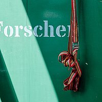 Detailaufnahme: an einer grünen Wand mit abblätternder Farbe baumelt von der leeren, orangenen Halterung eines Verbandkastens ein Spanngurt. Auf der Wand steht: Forschen