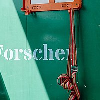 Detailaufnahme: an einer grünen Wand mit abblätternder Farbe baumelt von der leeren, orangenen Halterung eines Verbandkastens ein Spanngurt. Auf der Wand steht: Forschen