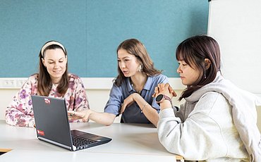 Drei Studierende sitzen am Tisch im Seminarraum und schauen in eine Präsentation auf dem Laptop.