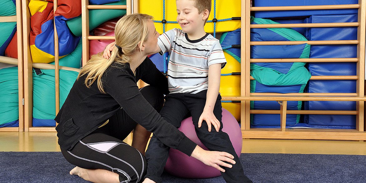 Eine Ergotherapeutin oder Physiotherapeutin zeigt einem auf einem Gymnastikball sitzenden kleinen Jungen Übungen.