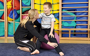 Eine Ergotherapeutin oder Physiotherapeutin zeigt einem auf einem Gymnastikball sitzenden kleinen Jungen Übungen.
