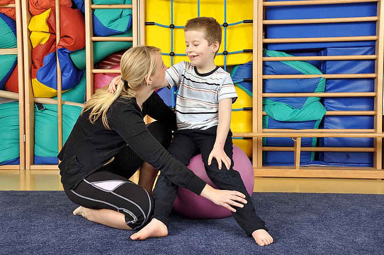 Eine Ergotherapeutin oder Physiotherapeutin zeigt einem auf einem Gymnastikball sitzenden kleinen Jungen Übungen. 