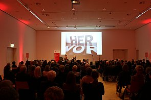 Blick von hinten auf das Publikum der Preisverleihung und eine Folie von Barbara Köhler, auf der steht "HERVOR" 