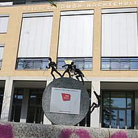Foto der Skulptur vor der Hochschule. Sie besteht aus einem halbrunden Stein. Oben stehen drei Figuren, die an einem Seil ziehen. Eine vierte Figur klettert an diesem Seil hoch. 