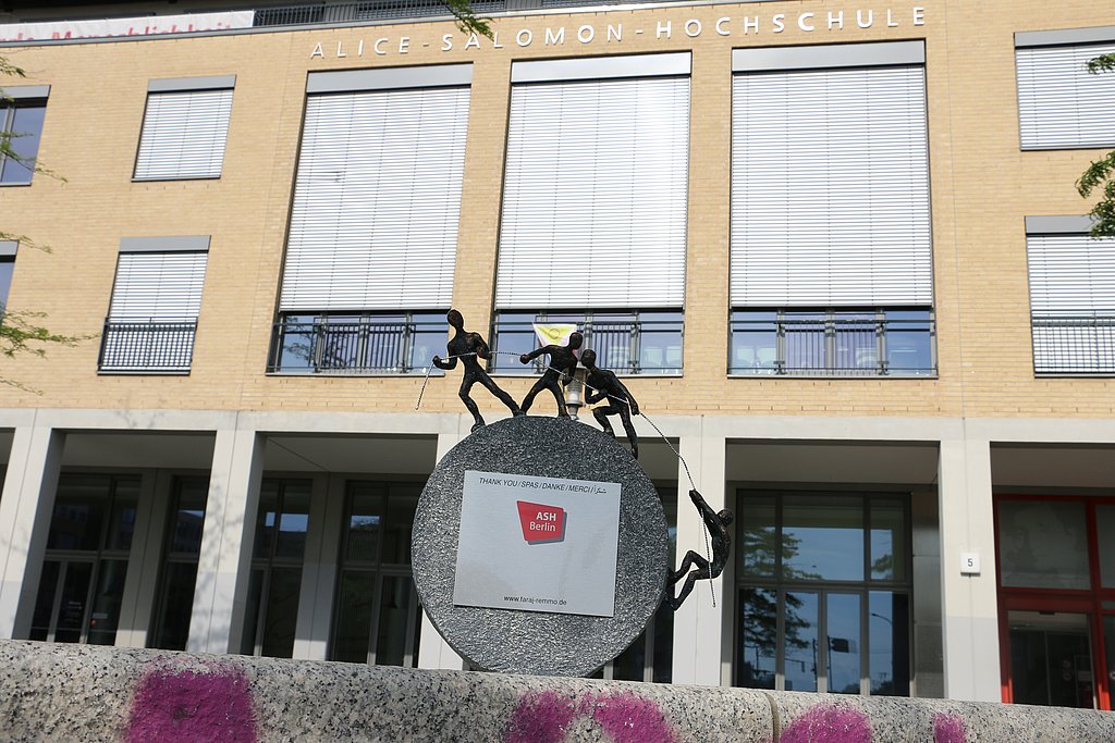 Foto der Skulptur vor der Hochschule. Sie besteht aus einem halbrunden Stein. Oben stehen drei Figuren, die an einem Seil ziehen. Eine vierte Figur klettert an diesem Seil hoch. 