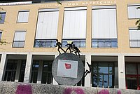 Photo of the sculpture in front of the university. It consists of a semicircular stone. At the top are three figures pulling on a rope. A fourth figure is climbing up this rope.