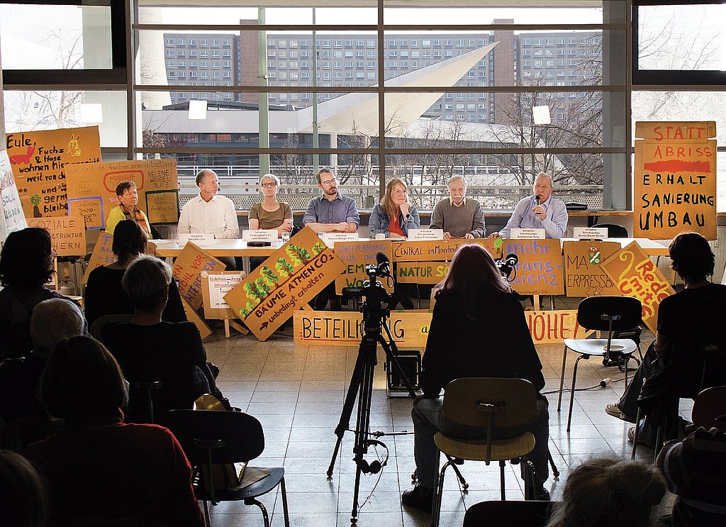 Das Bild zeigt eine Tagung der Anwohner*inneninitiative Marzahn-Hellersdorf. Mehrere Personen sitzen an einem langen Tisch und sprechen in ein Mikrofon, rund um sie herum sind Protestschilder aufgestellt. Das Foto ist mit Blick auf das Podium gemacht, mehrere Personen im Publikum sind von hinten erkennbar. Hinter dem Podium fällt der Blick auf Plattenbauten.