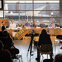 Das Bild zeigt eine Tagung der Anwohner*inneninitiative Marzahn-Hellersdorf. Mehrere Personen sitzen an einem langen Tisch und sprechen in ein Mikrofon, rund um sie herum sind Protestschilder aufgestellt. Das Foto ist mit Blick auf das Podium gemacht, mehrere Personen im Publikum sind von hinten erkennbar. Hinter dem Podium fällt der Blick auf Plattenbauten.
