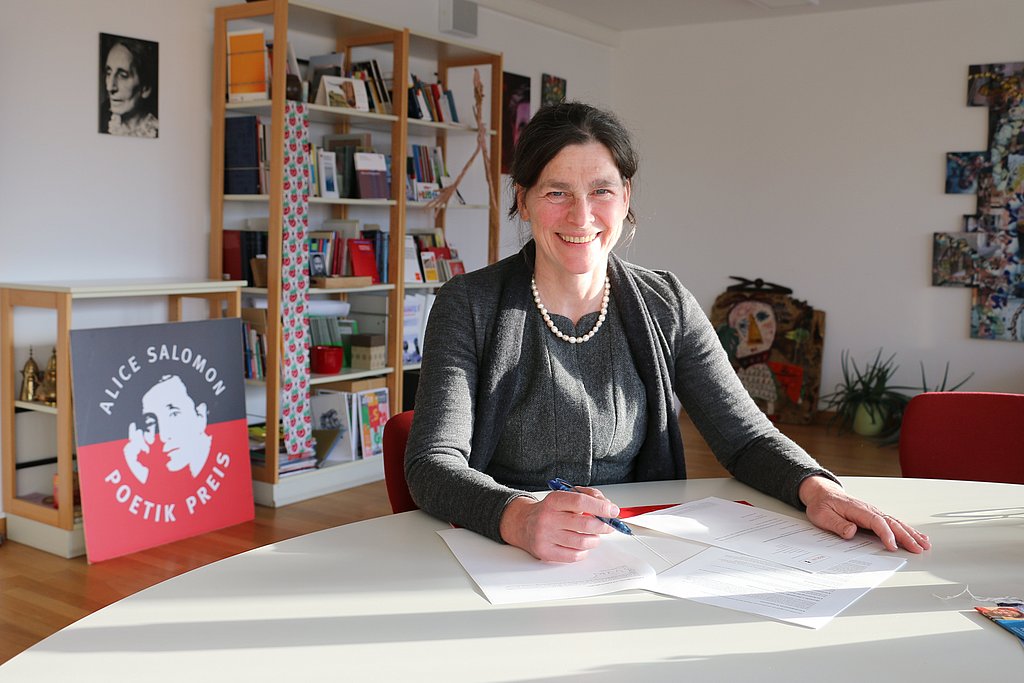 Auf dem Foto ist Bettina Völter, die Rektorin der ASH Berlin zu sehen. Sie sitzt an einem Tisch in ihrem Büro und schaut in die Kamera. Vor ihr liegt die neue Grundordnung der Hochschule, die sie soeben unterzeichnet hat. 
