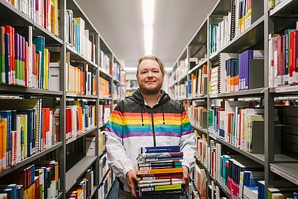 Eine Person steht in einem Gang in der Bibliothek, der Kamera zugewandt, hält einen Stapel Bücher in der Hand, trägt einen Kapuzenpullover mit Regenbogenmuster.
