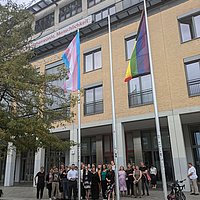 Auf dem Foto ist eine große Gruppe von Personen vor der ASH Berlin zu sehen, außerdem die Progressiveflag und die Transprideflag, die an den Fahnenmasten wehen. 