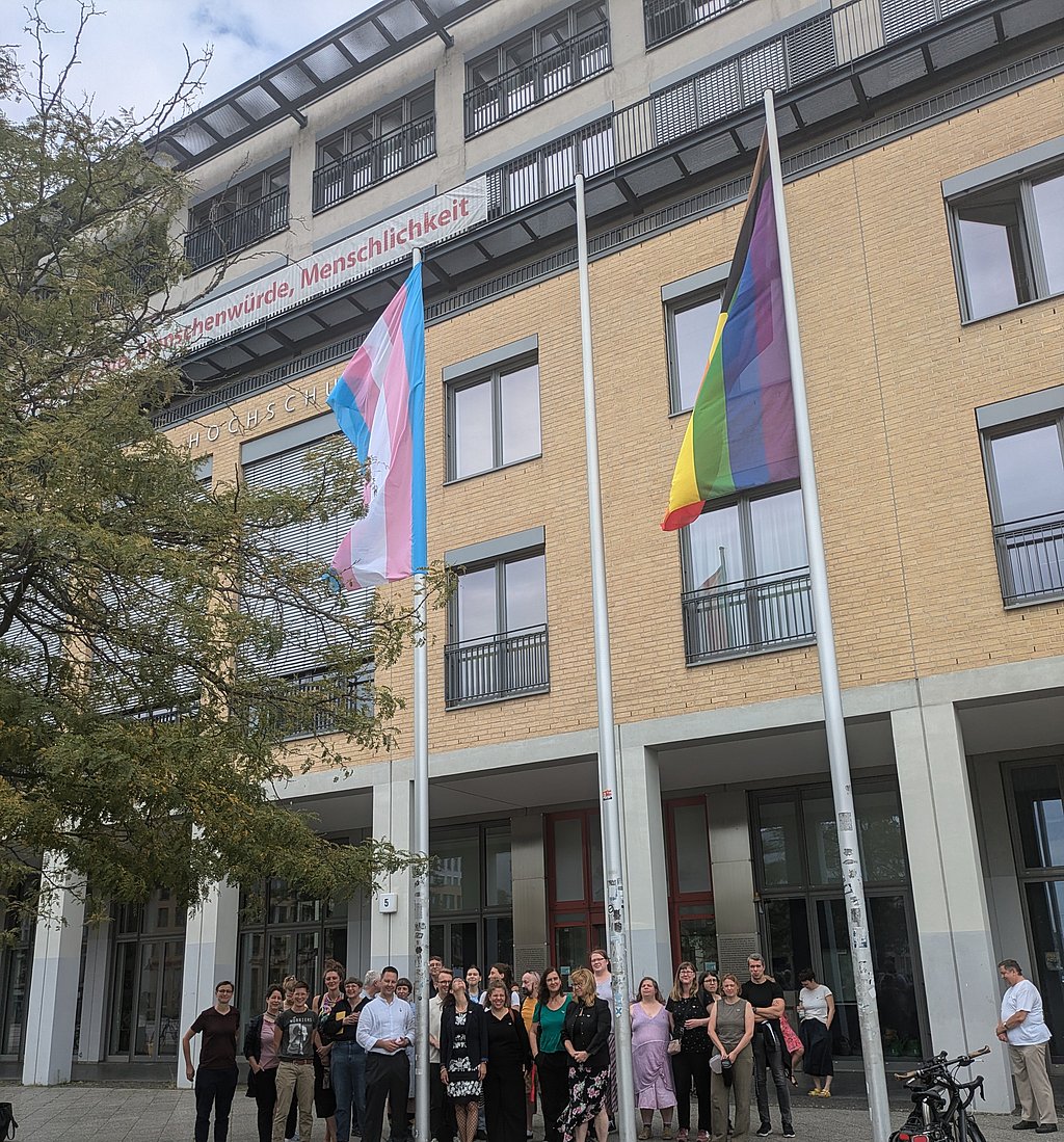 Auf dem Foto ist eine große Gruppe von Personen vor der ASH Berlin zu sehen, außerdem die Progressiveflag und die Transprideflag, die an den Fahnenmasten wehen. 