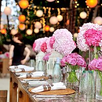 Das Bild zeigt einen festlichen gedeckten Tisch mit rosa Blumensträußen, im Hintergund sind in Bäumen hängende Lampions zu sehen.