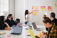 Menschen sitzen an einem Tisch und besprechen ein Thema. Auf dem Tisch stehen Laptops. Eine Person steht am Whiteboard.