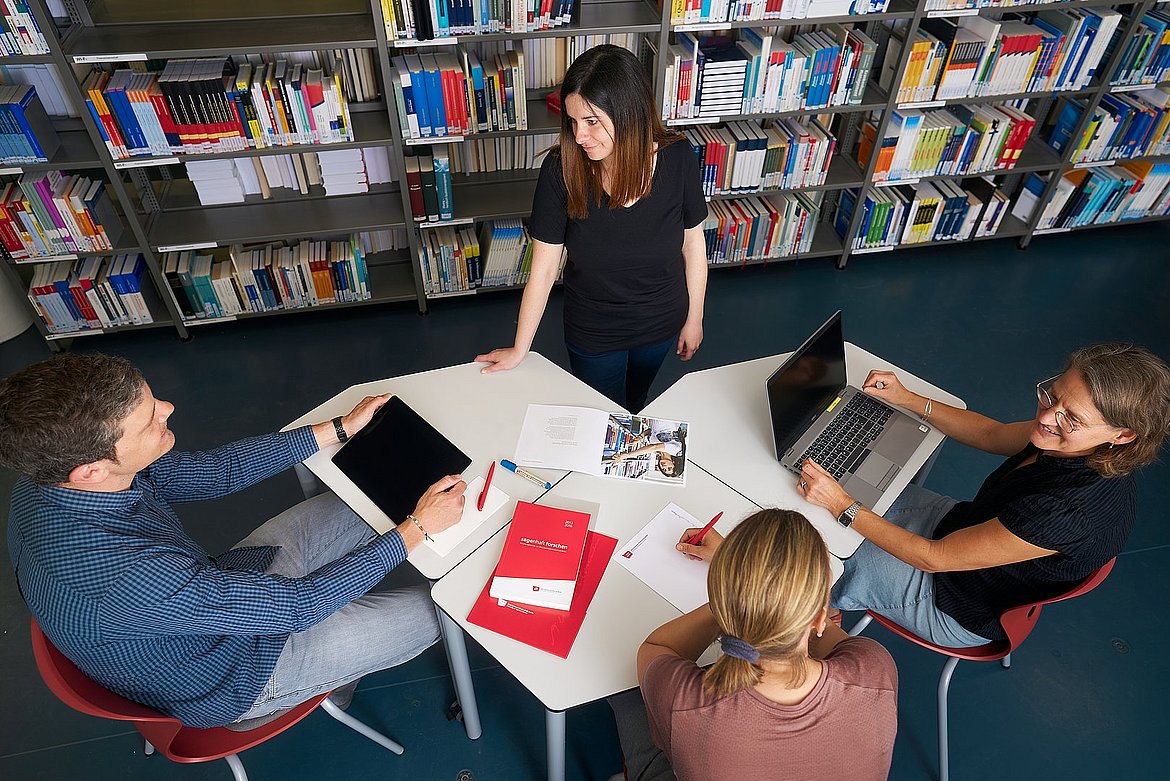 Eine dargestellte Schulungssituation in der ASH-Bibliothek mit vier Personen. Drei Personen sitzen am Tisch und eine Person steht.
