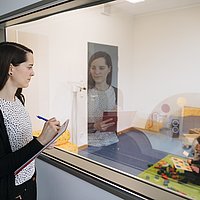 Eine Wissenschaftlerin beobachtet durch einen Einwegspiegel die Interaktion eines Kleinkindes mit seiner Mutter. 