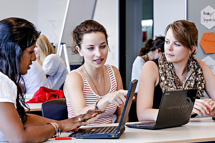 drei Studierende sitzen vor ihren Laptops, eine Studentin zeigt der anderen etwas