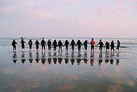 Mehrere Menschen stehen am Meer mit den Füßen im Wasser und halten sich dabei an den Händen. 