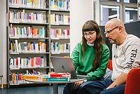 Zwei Studierende sitzen in der ASH-Bibliothek in den Laptop schauend. Im Hintergrund ein Regal mit Büchern.