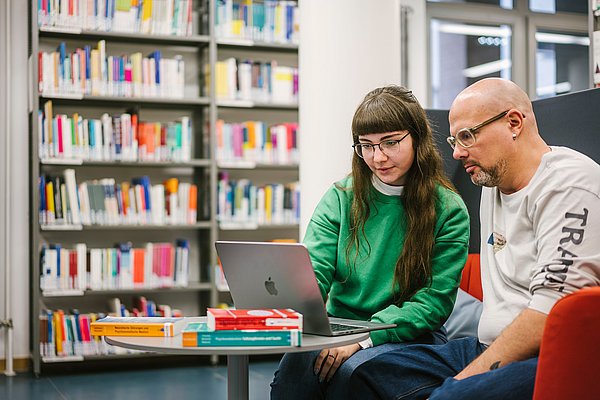 Zwei Studierende sitzen in der ASH-Bibliothek in den Laptop schauend. Im Hintergrund ein Regal mit Büchern.