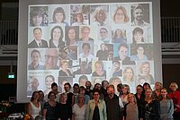 Die Absolventi*innen des Igo-Studiengangs stehen zum Gruppenbild im Audimax, hinter ihnen eine Leinwand mit ihren Portraitfotos