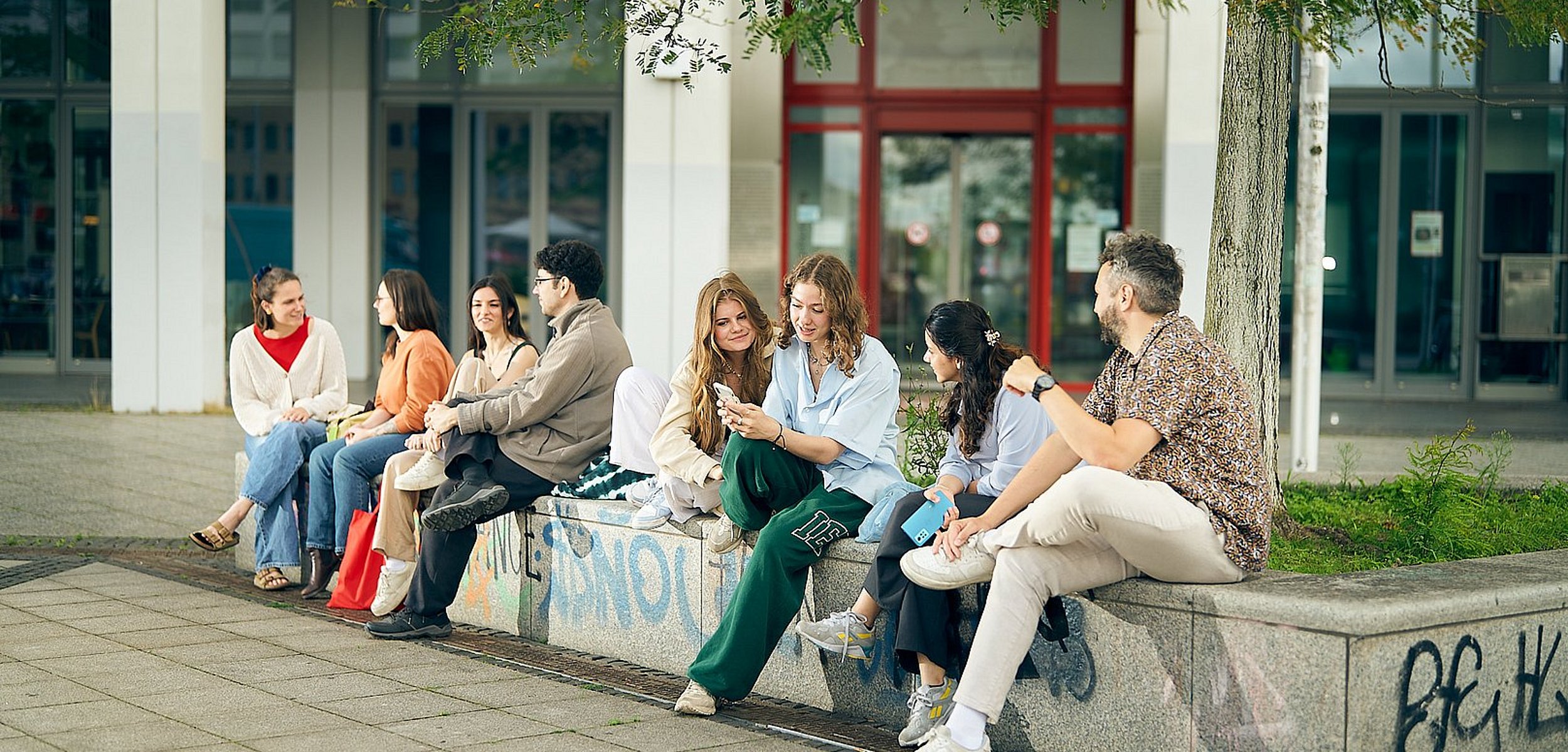 Gruppenfoto von Studierenden der ASH Berlin die vor der Hochschule sitzen und miteinander reden.